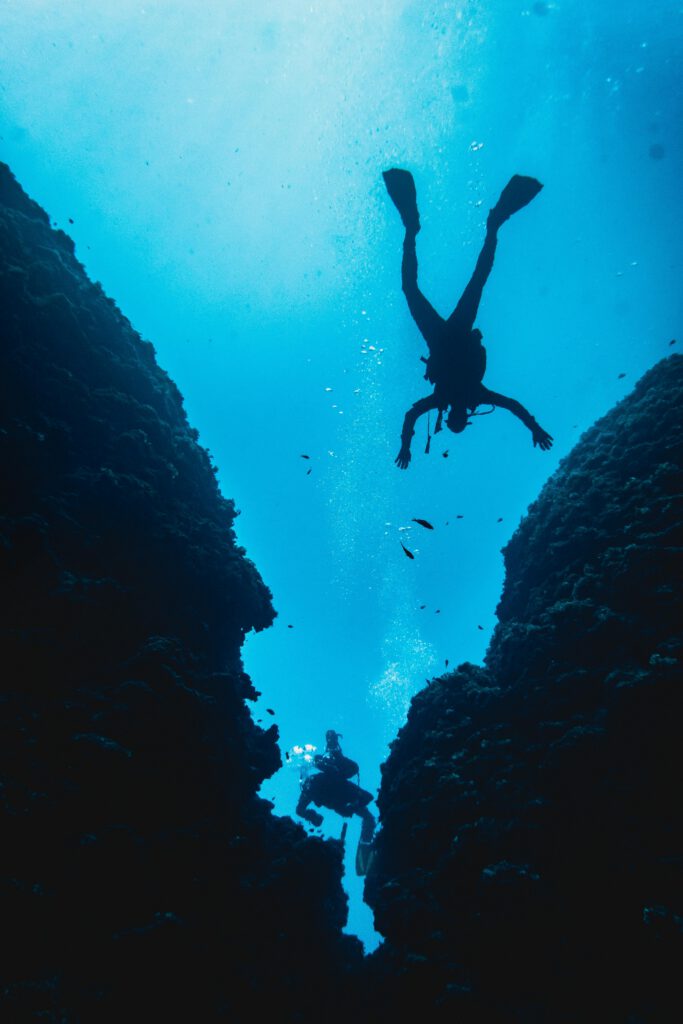 From below of anonymous male tourists in protective outfit and flippers exploring blue ocean water near big reefs during trip